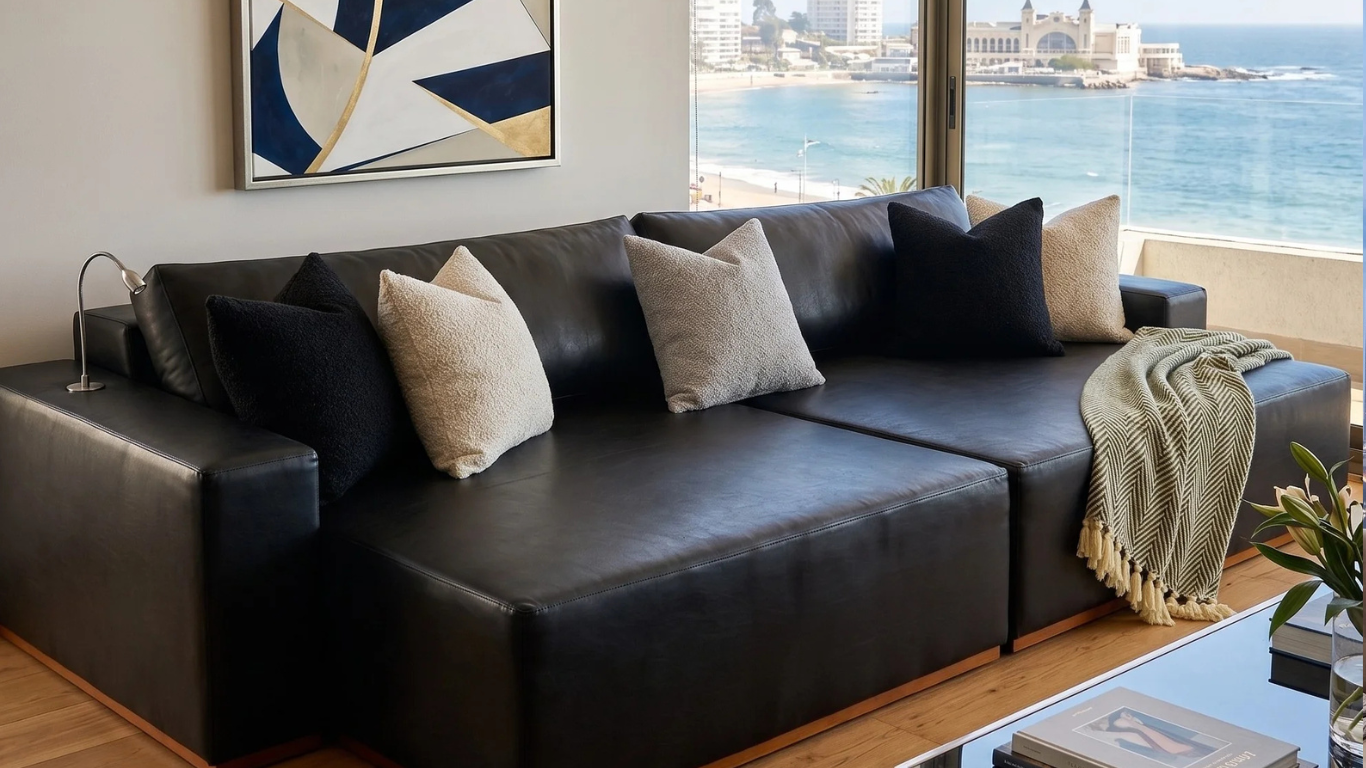 Departamento moderno con vista a la costa, sofá modular de cuero negro, cojines texturizados en blanco y negro, manta verde con flecos y un gran cuadro geométrico azul y dorado.