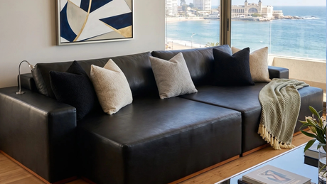 Departamento moderno con vista a la costa, sofá modular de cuero negro, cojines texturizados en blanco y negro, manta verde con flecos y un gran cuadro geométrico azul y dorado.