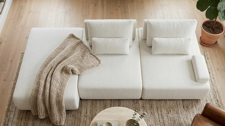 Plano cenital de living acogedor con sofá modular Alba blanco, decorado con una gruesa manta tejida color arena drapeada en el asiento, sobre una alfombra rústica de yute y suelo de madera clara.