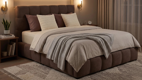 Dormitorio acogedor e iluminado con cama capitoné marrón completamente vestida, cojines texturizados y manta de punto gris.