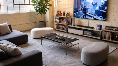 Sala recreativa Espacio de entretenimiento con alfombra gris clara, mesa de centro metálica negra, dos poufs curvos (uno blanco y uno gris) y televisor.