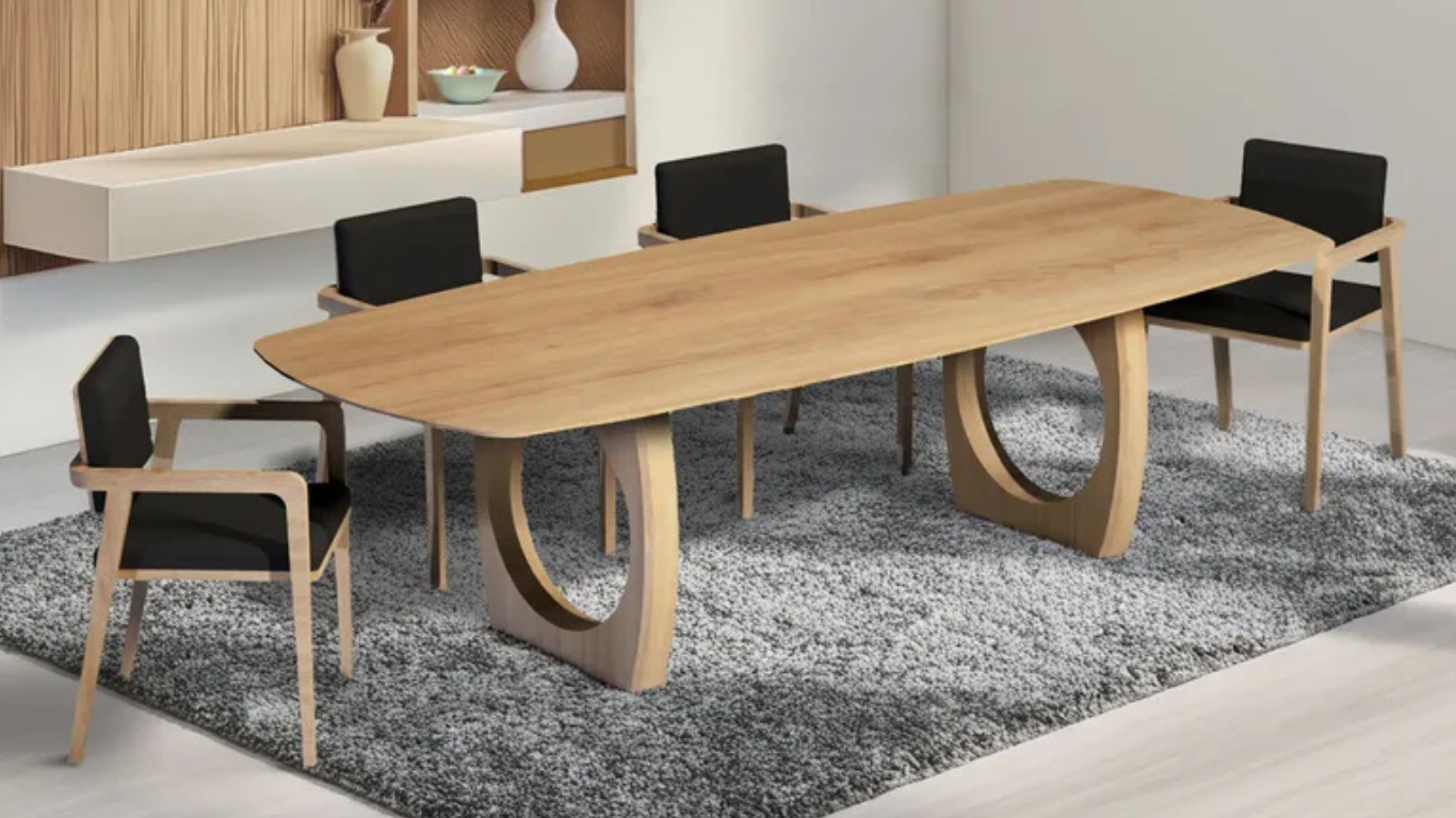 Comedor moderno con mesa rectangular de madera clara y base escultórica circular, acompañado de sillas con estructura de madera y cojines negros, sobre alfombra gris de textura suave.